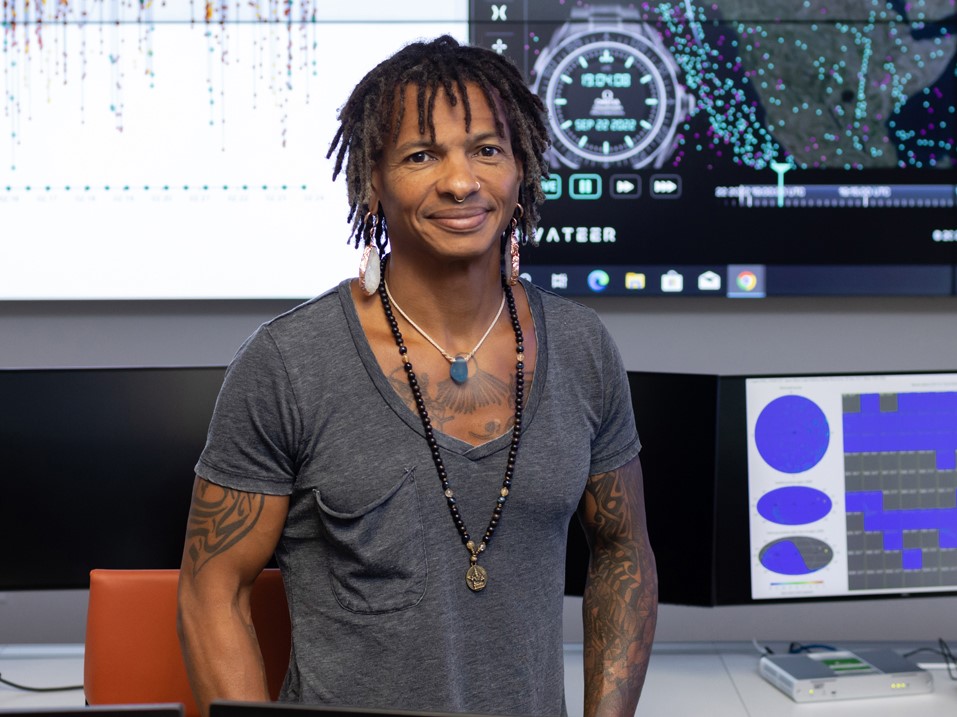 Moriba Jah smiling in front of computer screens with research displayed